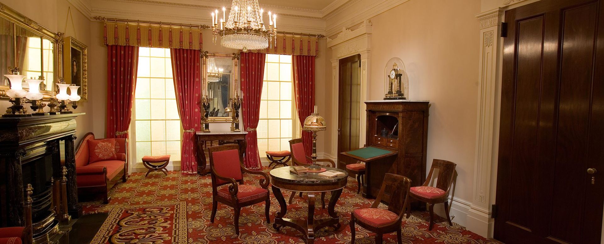 A wide view of the Greek Revival Parlor. A fireplace and mantel, table with four chairs, chandelier, and secretary are seen