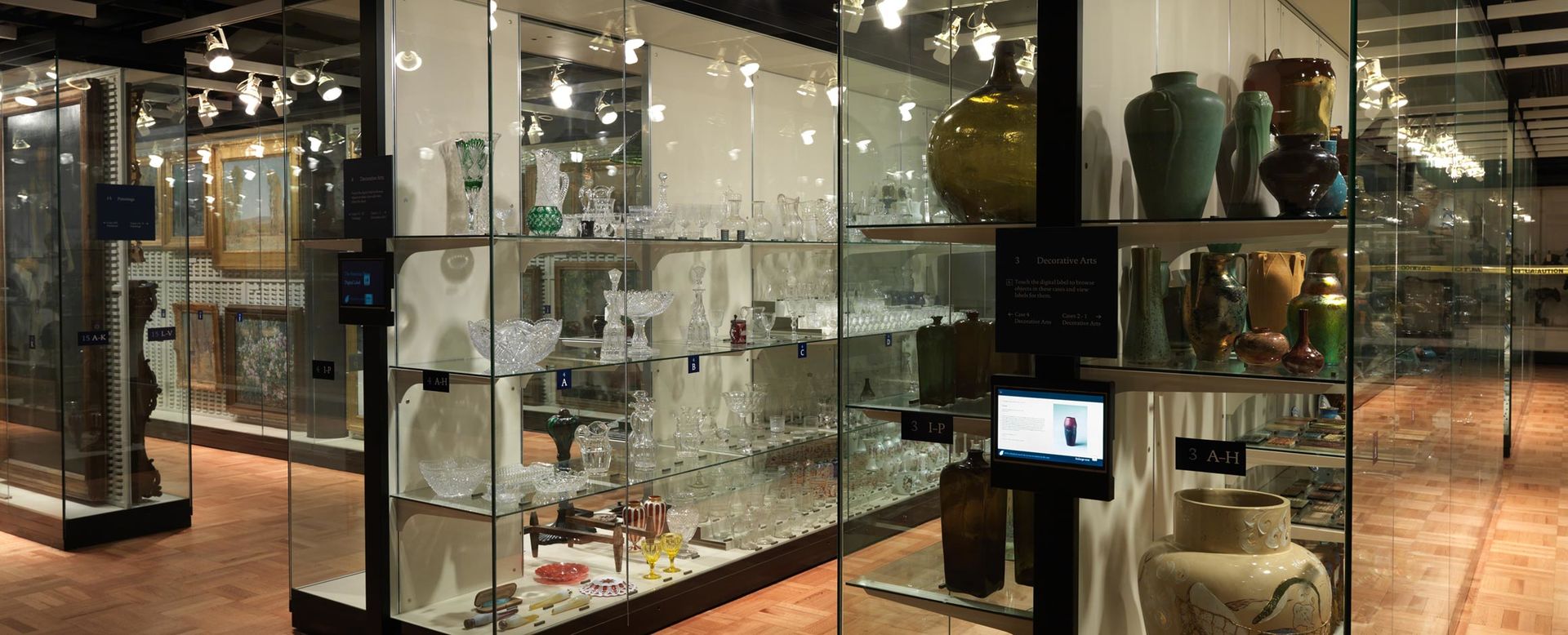 A gallery filled with glass-fronted shelving filled with art objects from the American Wing; touch screens are mounted along side all the cases