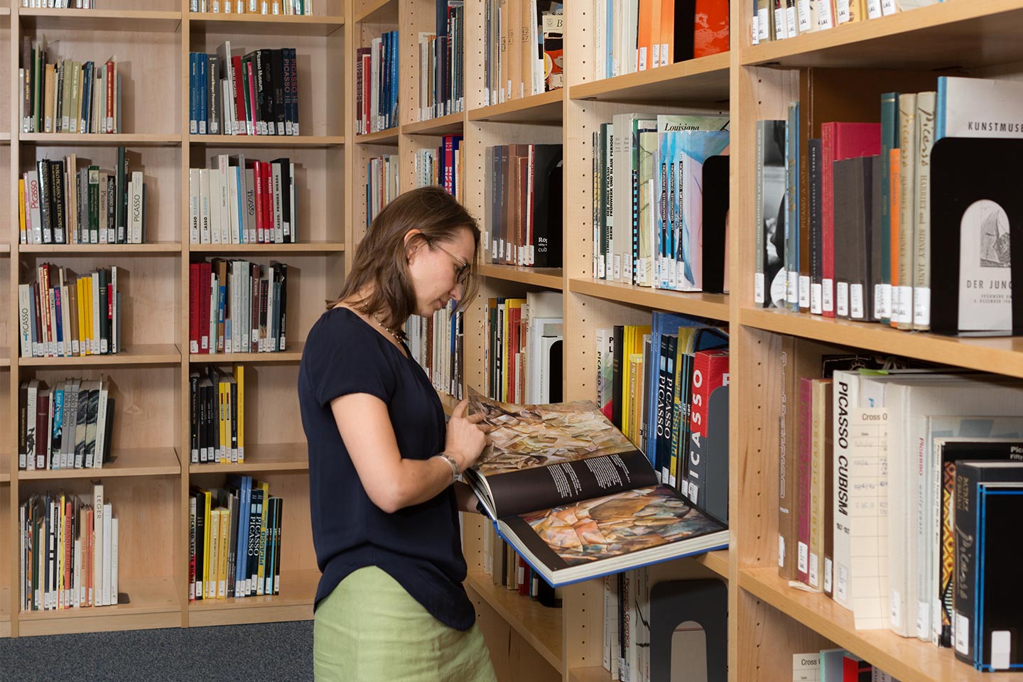 A woman stands with an open book in her hands in the corner of a wooden bookshelf