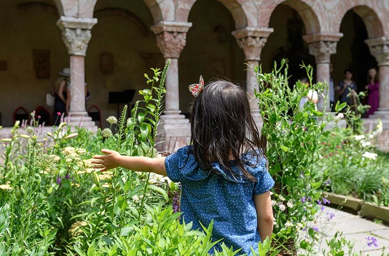 a young girl with her back turned to the camera stands in front of a garden bed in a medieval cloister.
