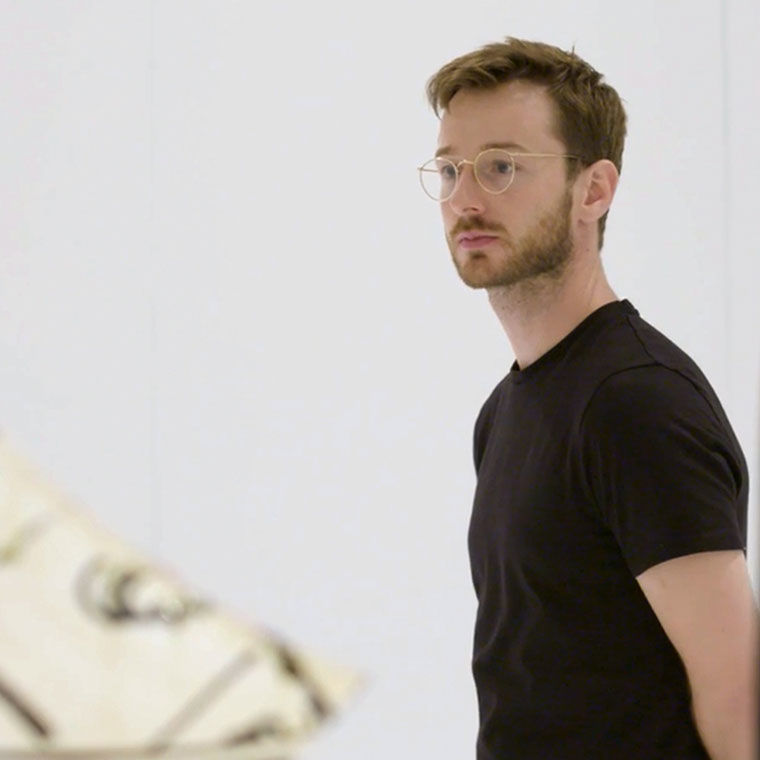 A blonde-haired man with glasses, dressed in black, stands in a gallery with an out of focus sculpture in the background