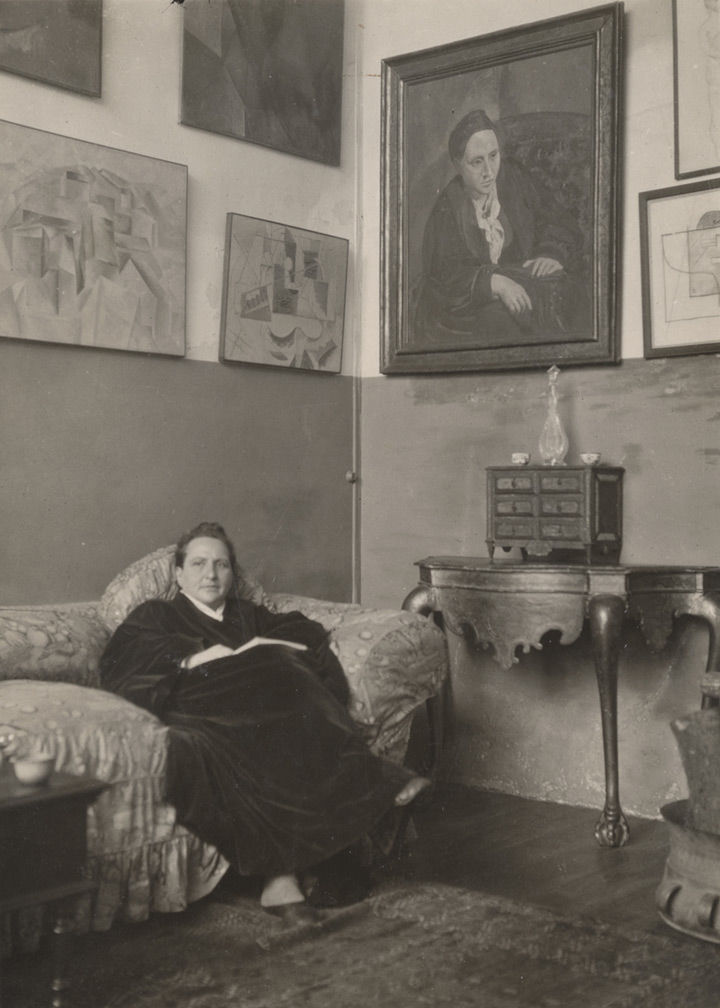 A black-and-white photo of Gertrude Stein seated in her salon, beneath a portrait of her by Pablo Picasso