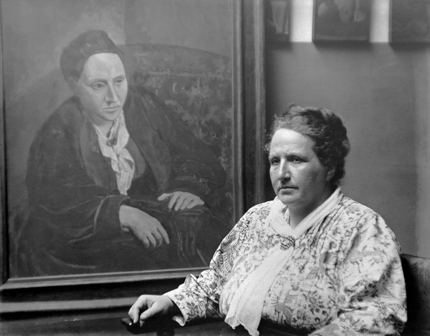 Black and white photograph of a woman wearing a white floral pattern outfit on the right with a potrait in the background done by Picasso of her. 