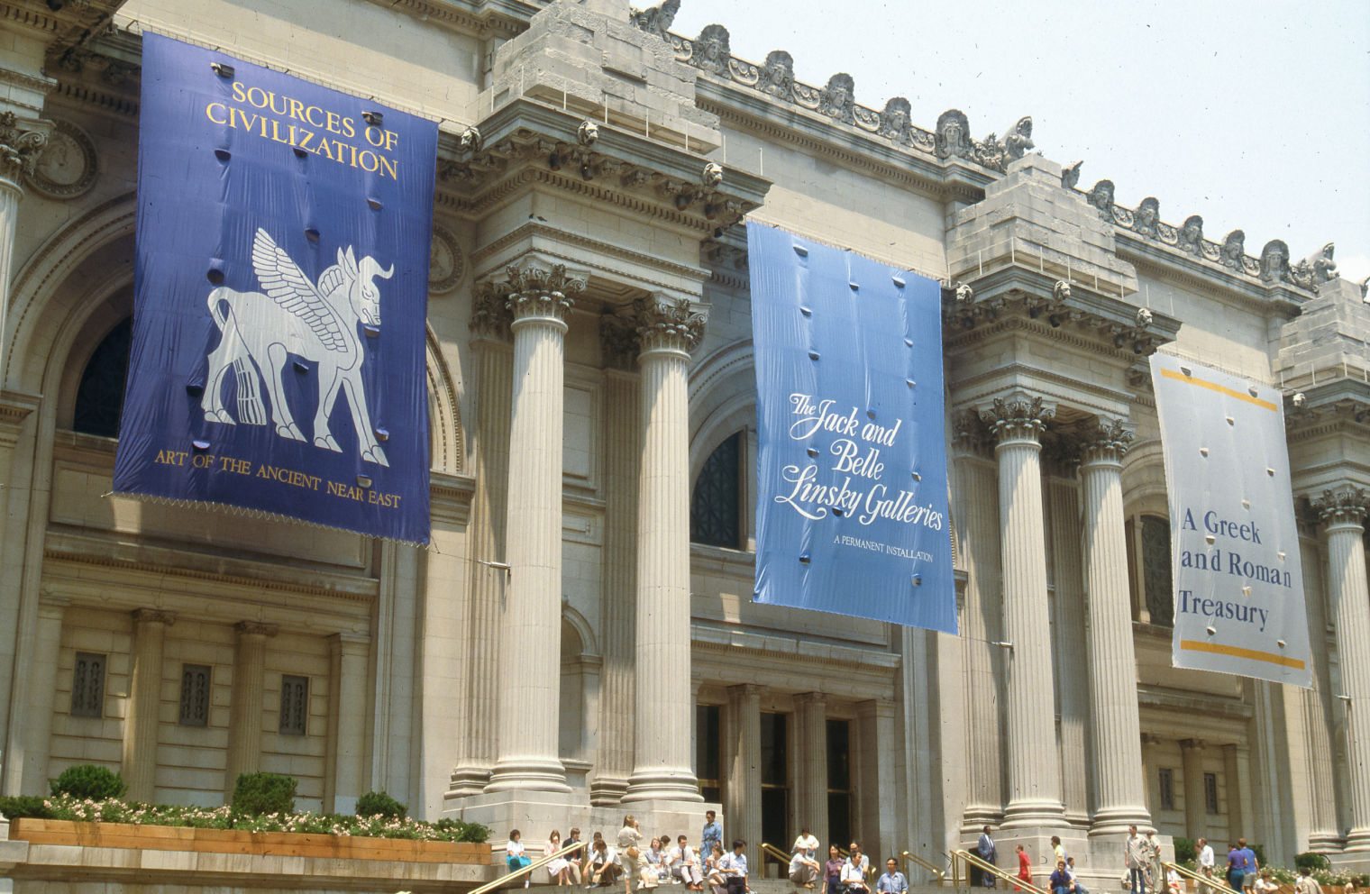 Entrance of the Metropolitan Museum of Art with people showing, left to right, banners for  “The Ancient Near Eastern Art Gallery,” “The Jack and Belle Linsky Collection,” “Greek and Roman Art Gallery”