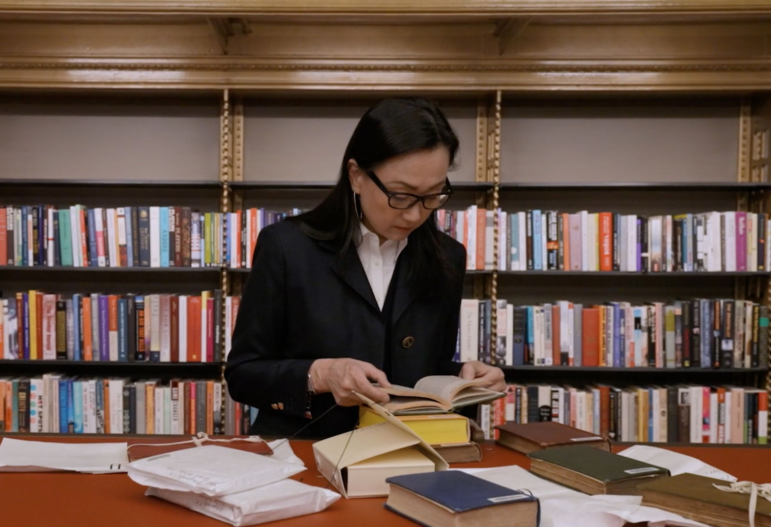 The Novelist Min Jin Lee peruses through an assortment of books atop a red-orange tabletop surface, set against a backdrop of book shelves in a New York Public Library study room.