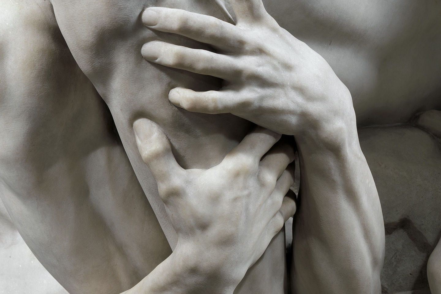 A close up view of a carved marble sculpture depicting muscular hands wrapping around and gripping a figure’s leg to the side of the knee cap.