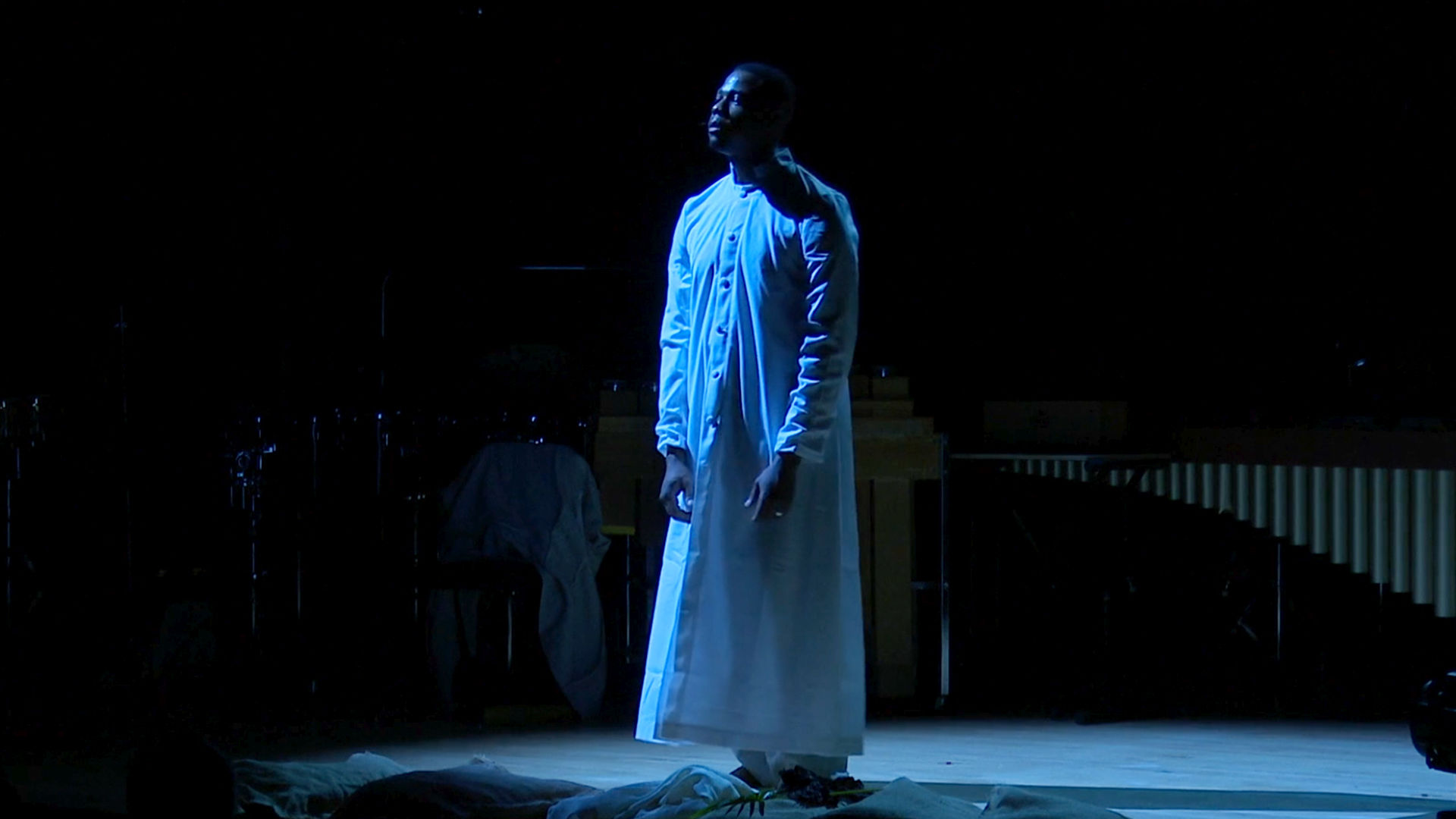 A dark skinned man wearing a long white button-down robe stands on a stage lit by a blue light.