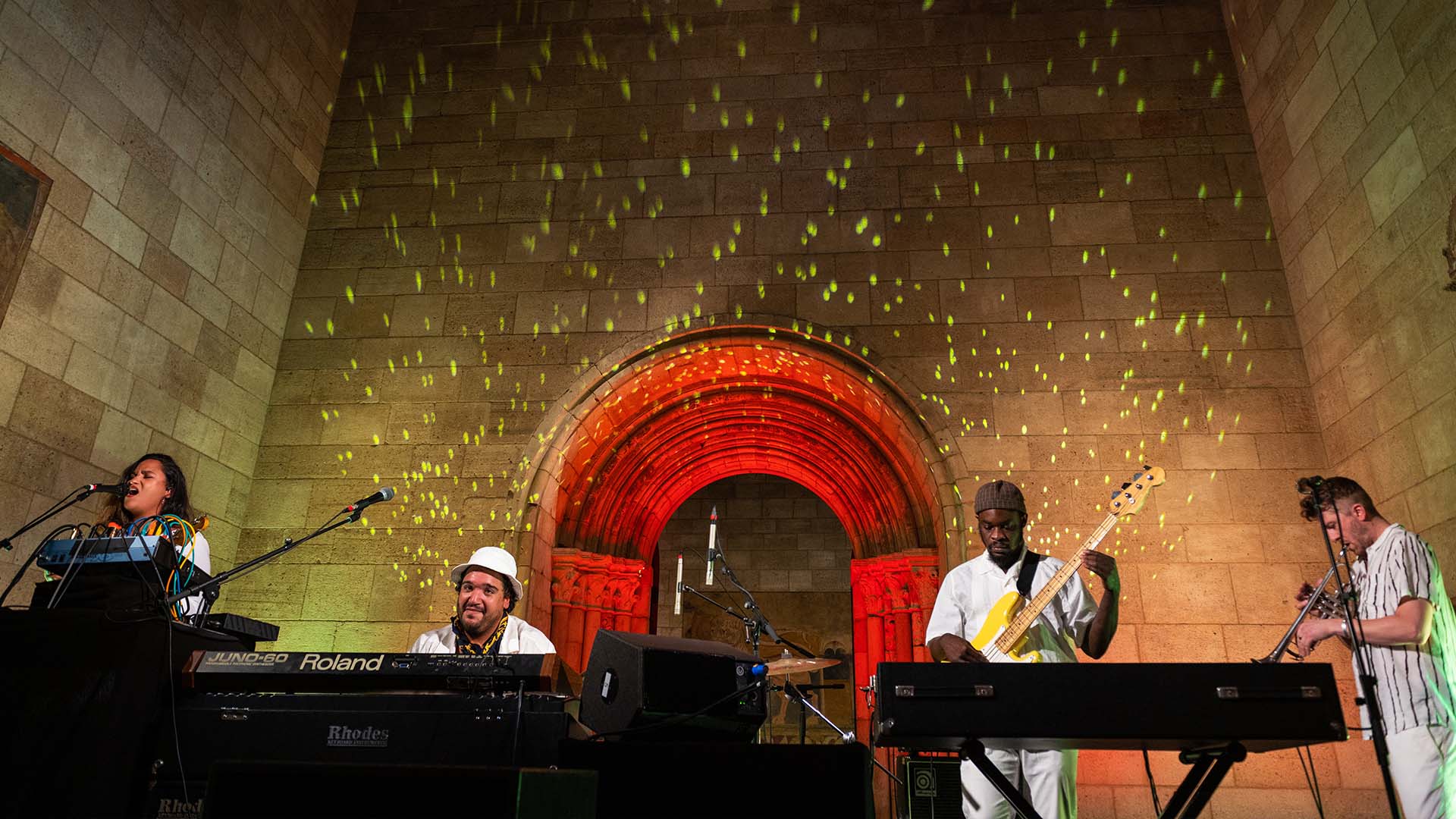 Four musicians performing at a live concert in front of an arched wall lit up with red and yellow lights.