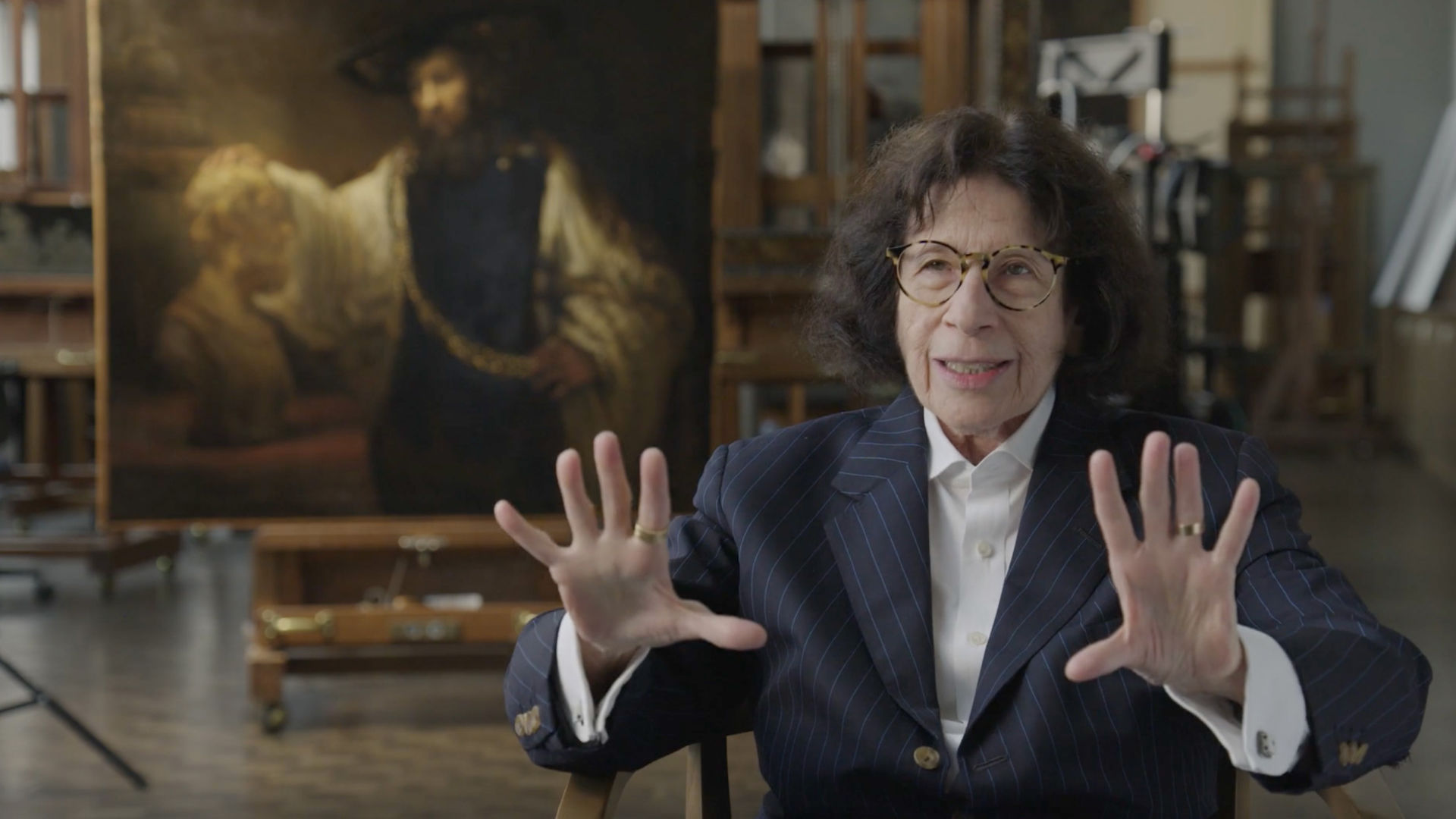 Writer Fran Lebowitz speaking and gesturing with her hands in The Met’s Paintings Conservation Lab in front of Rembrandt’s Aristotle with a Bust of Homer painting.