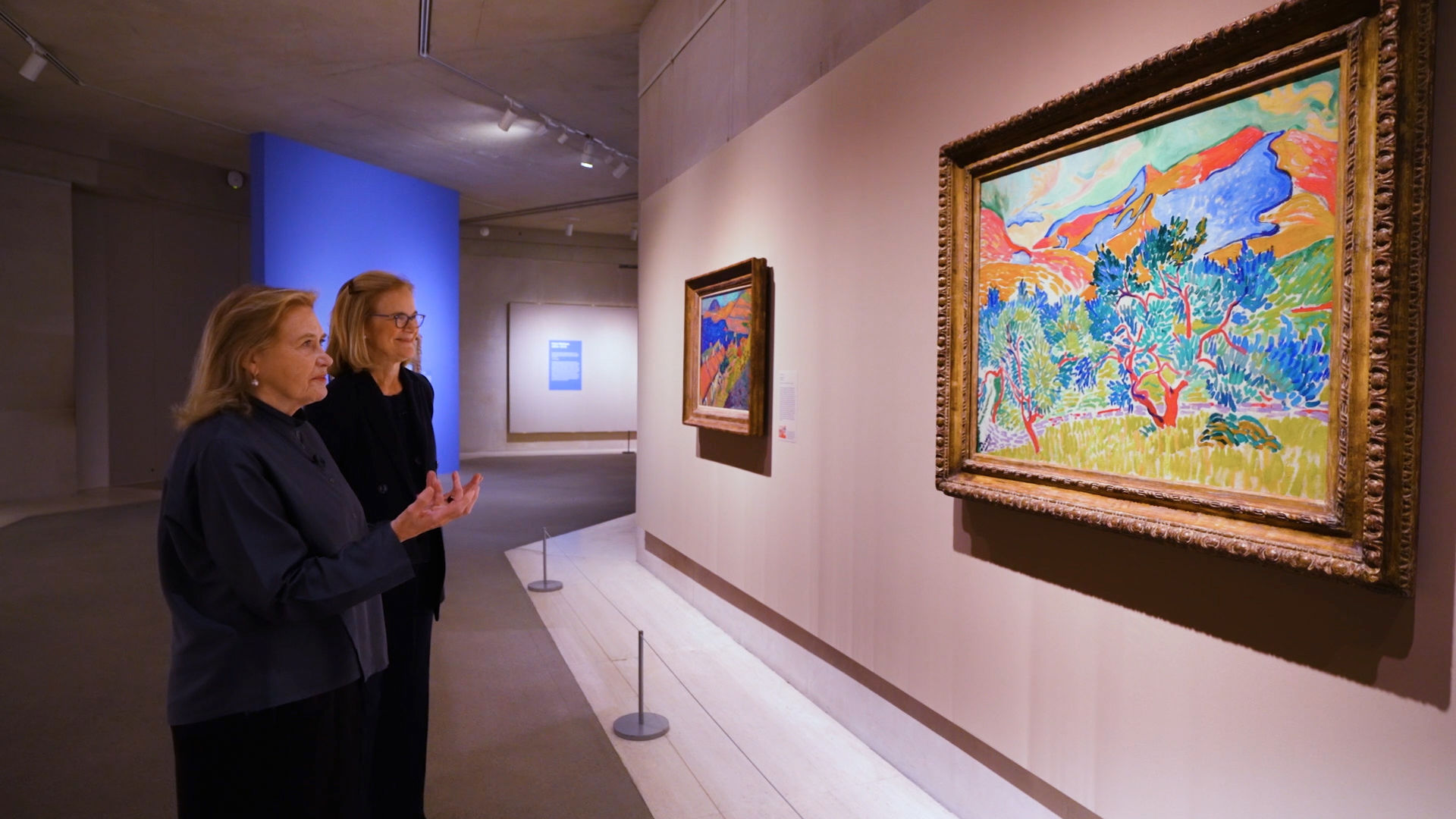 Two women in conversation with each other stand before a colorful landscape painting in a gold frame.