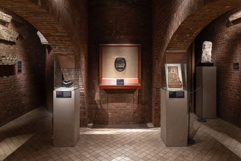 A gallery made of brick with two arches on either side. In the center, a mirrored oval mounted inside a glass case, on either side are two tall podiums with objects inside clear cases.