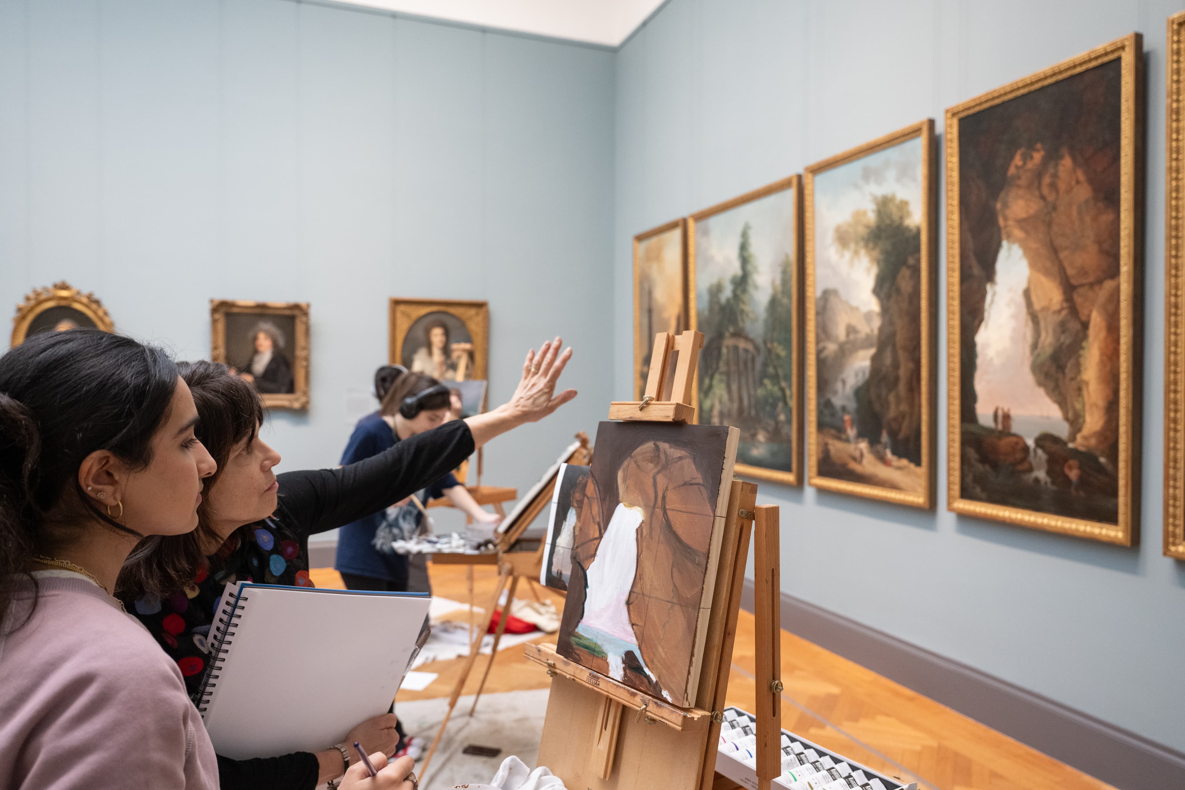 A teen views her in-progress painting with an educator who gestures towards her canvas. Her painting is setup on an easel and the artwork she is copying from hangs on the gallery wall.