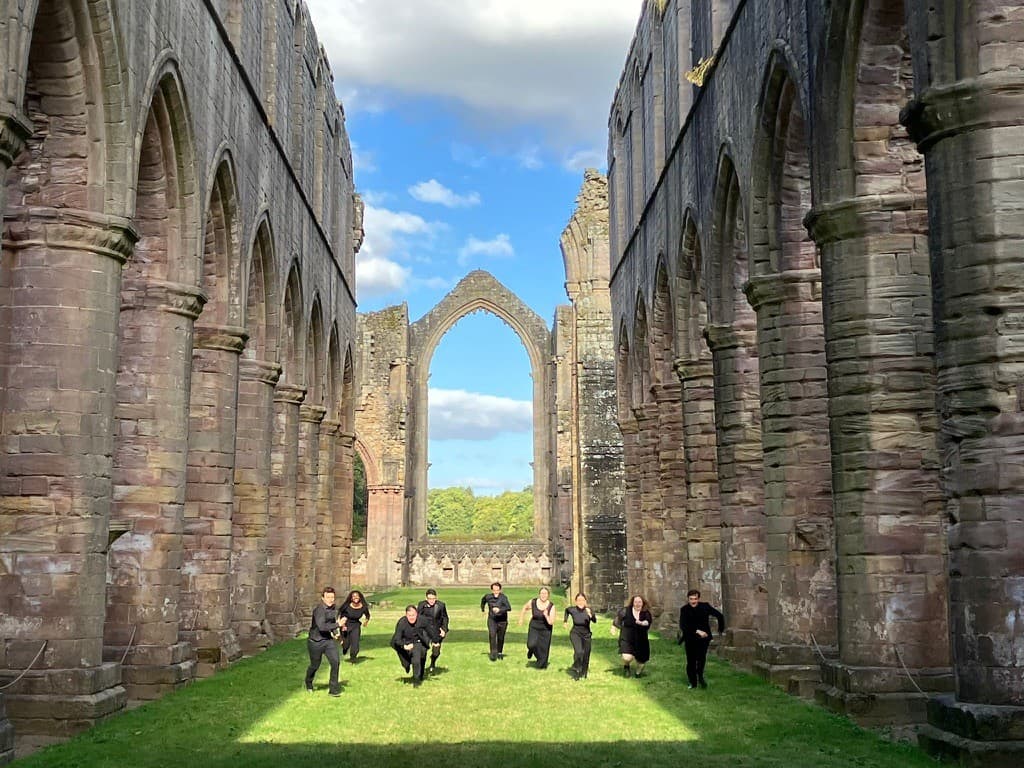 9 adults dressed in black standing in a grassy field between two aged corridors of archways