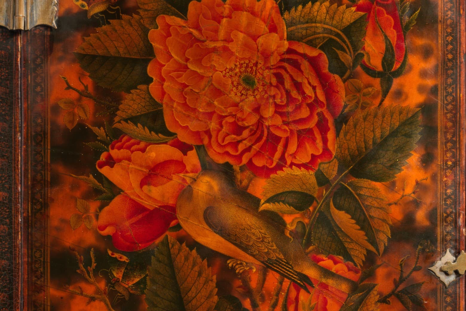 A mirror case decorated with flowers in hues of red and orange, with the figures of two people in the top corners, and a building in the bottom right corner.