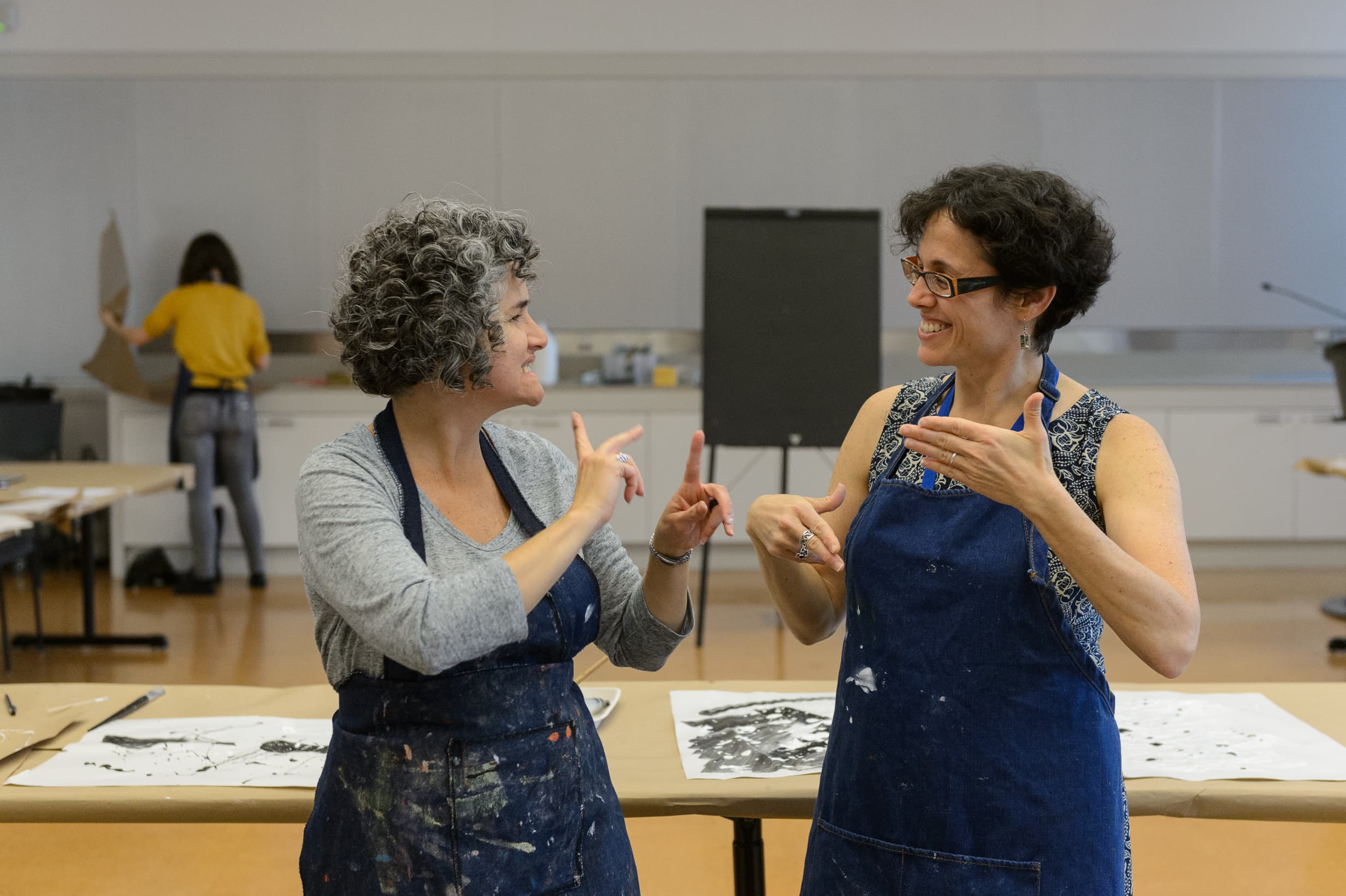 On the right, a Met educator with short black curly hair signs while in conversation. The person on the left has curly grey hair. Both are wearing blue denim smocks in an art studio.