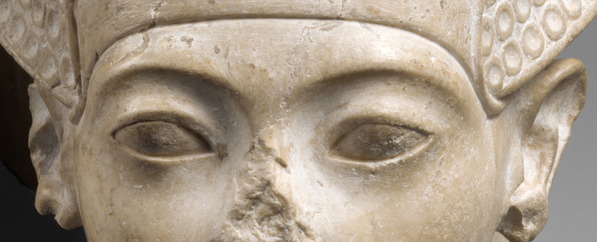 A close-up of a face, focused on the eyes, which are almond-shaped. Part of the area of the nose, which was broken away, is visible, Above the eyes is the band of a helmet-like crown.