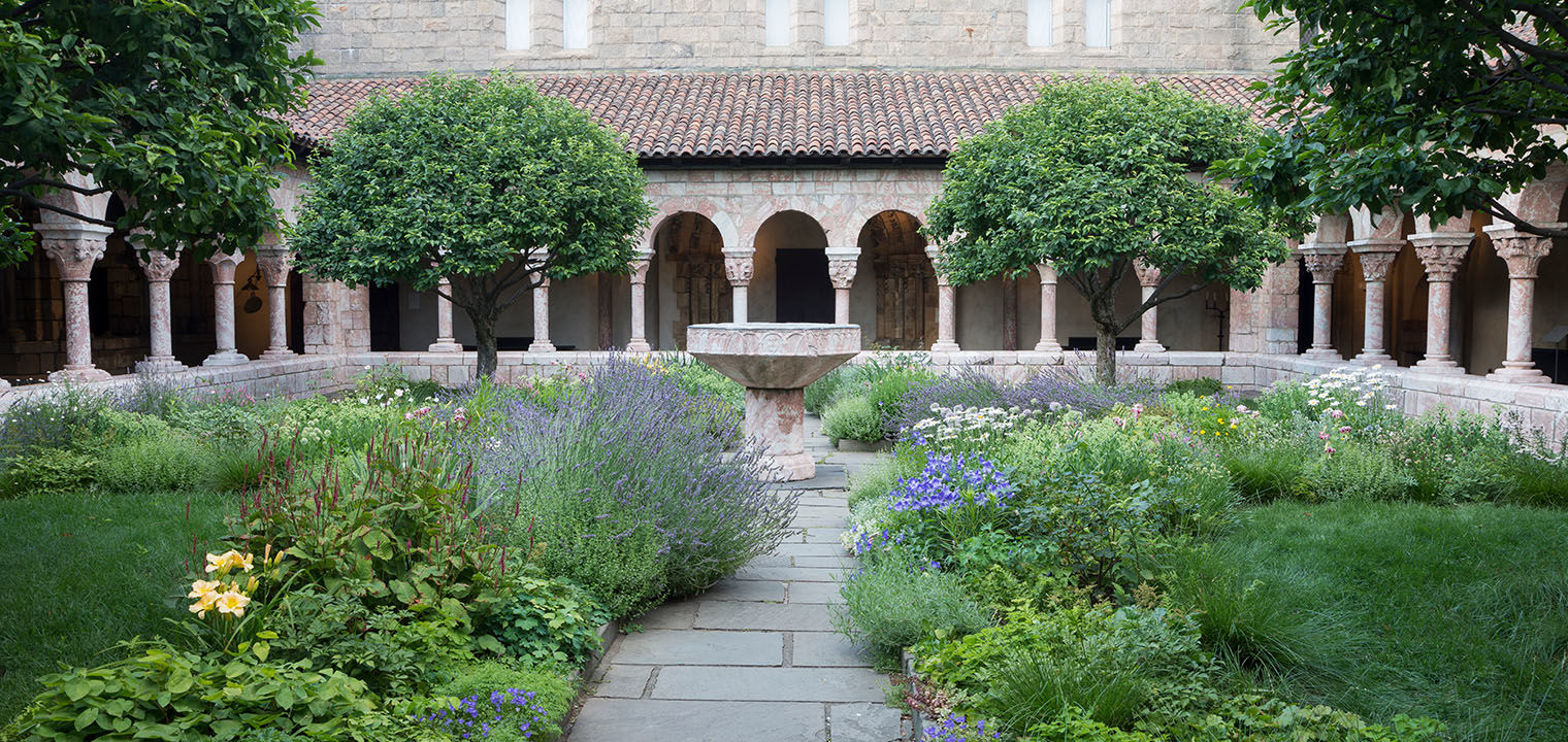 A garden courtyard surrounded by a arched colonnades; a central stone pathway with luscious plant beds encroaching on either side leads to a central stone fountain