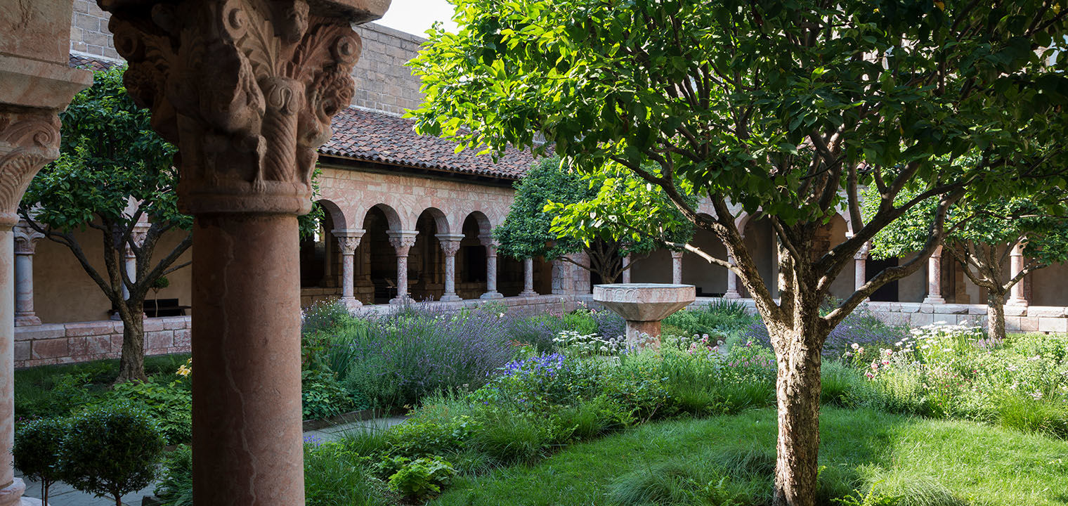 A romanesque column and tree flank and frame a view of a lush garden courtyard on a sunny day