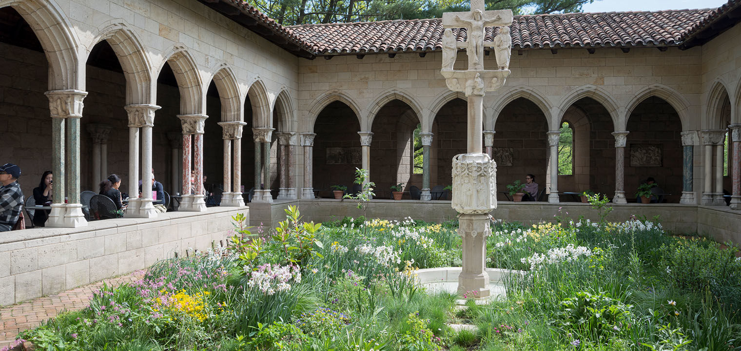 A garden courtyard consisting of a meadow of flowering plants and central stone statue; the courtyard in surrounded by a colonnaded passageway with visitors dining