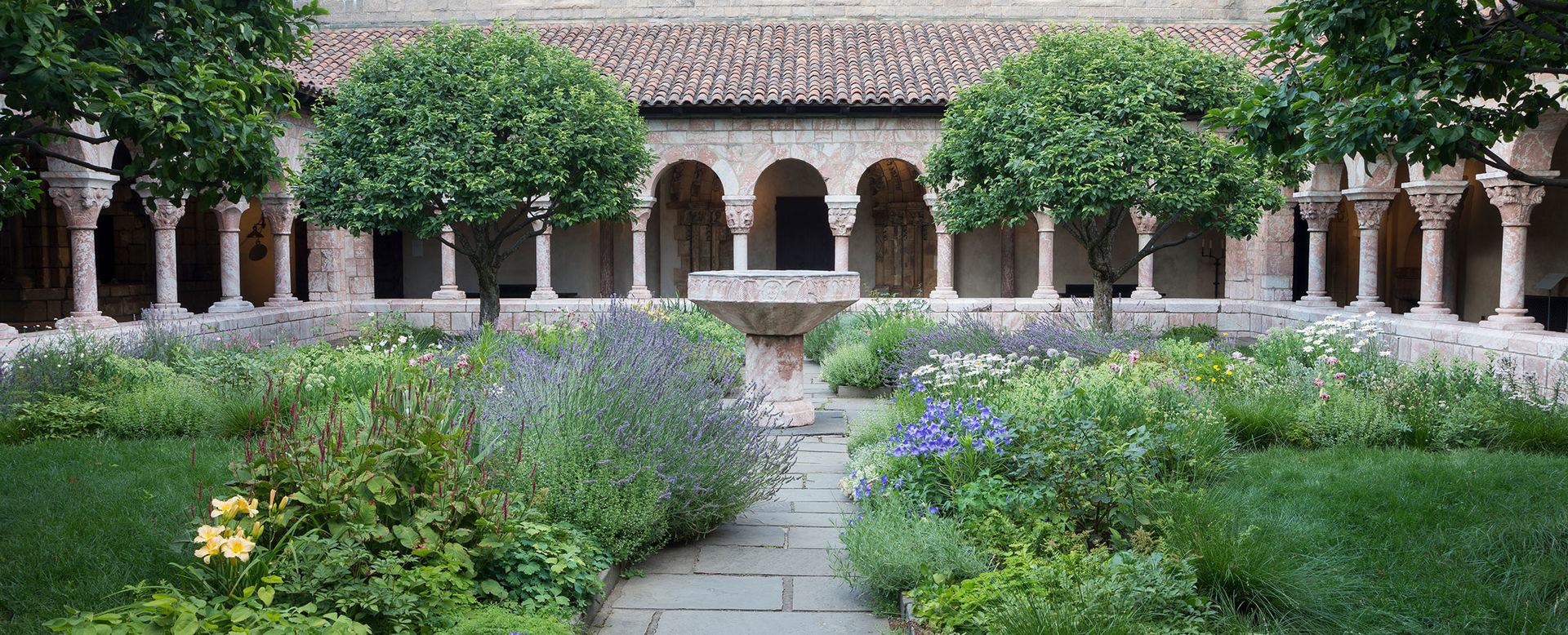 A garden courtyard surrounded by a arched colonnades; a central stone pathway with luscious plant beds encroaching on either side leads to a central stone fountain
