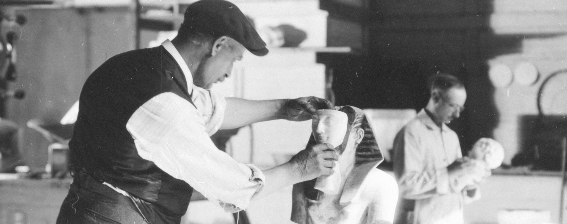 Detail of archival black-and-white photograph showing a man in a white shirt and black vest wearing a black cap fitting the face onto an Egyptian head
