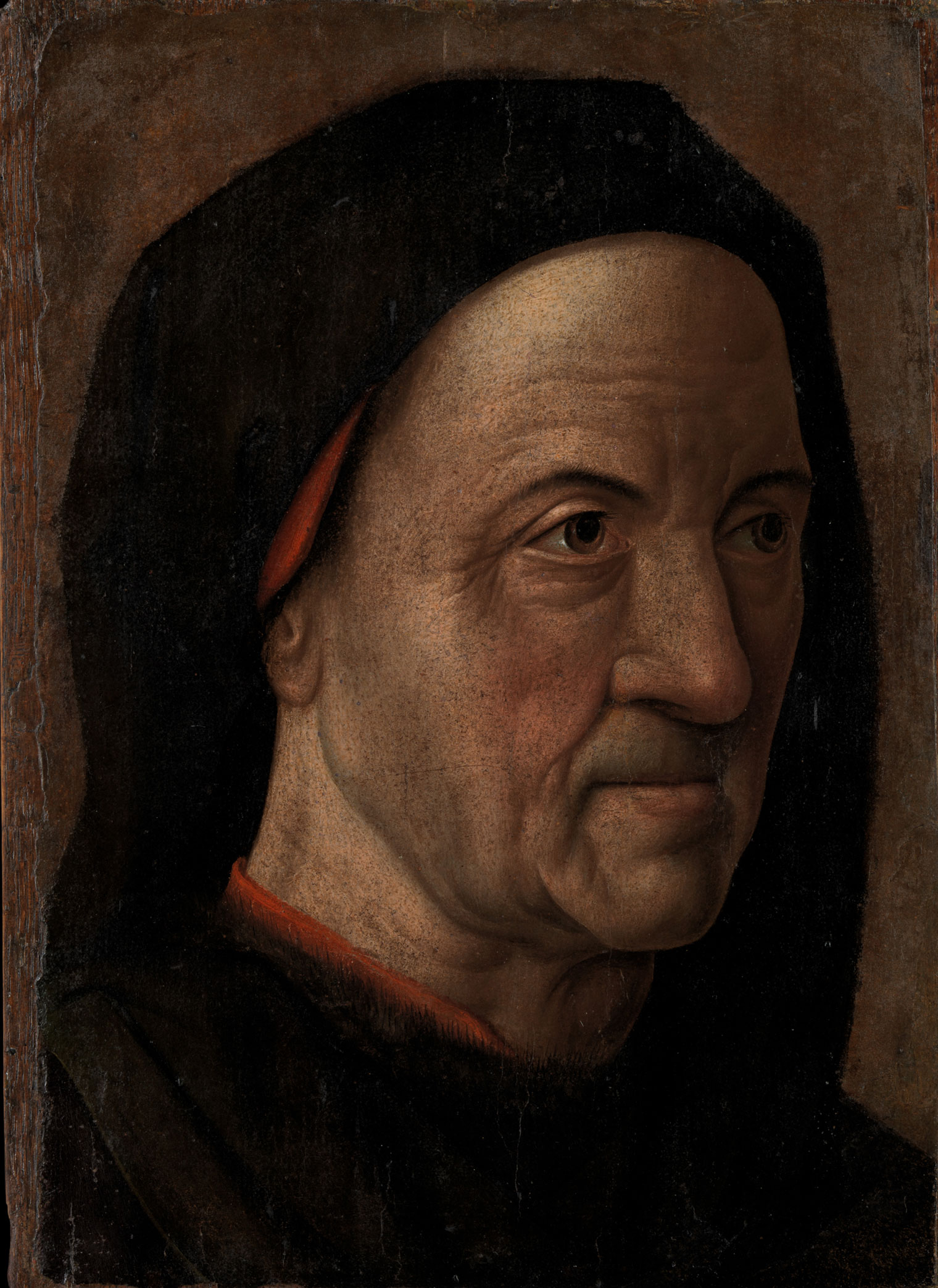 A detail of a portrait by Hugo van der Goes depicting a wrinkled old man wearing a black cloak with a red garment underneath in front of a uniform brown background.