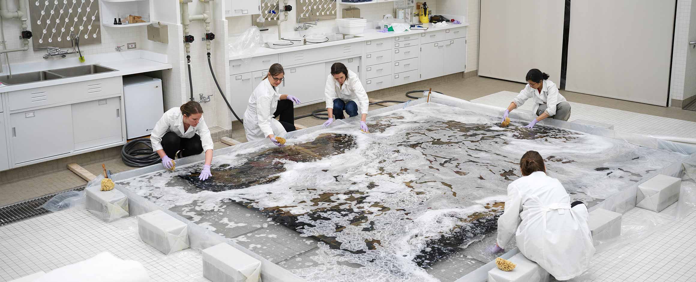 A group of textile conservators wearing white lab coats and purple gloves use sponges to wash a tapestry in their lab