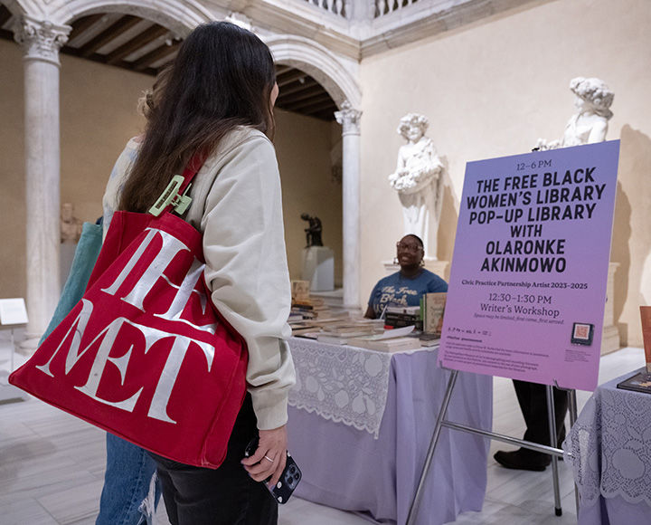In the sunlit Vélez Blanco Patio, two tables shrouded in a lavender-colored cloth with lace overlay are covered with book displays. A person is seated at the table to the left, smiling at two visitors perusing the installation. A sign to the right of the table advertises a pop-up for the Free Black Women's Library.