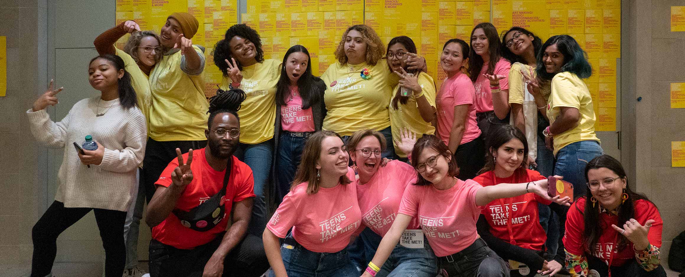 A group of teens are waving and making fun faces to the camera. They are wearing pink and yellow t-shirts, and look very happy.