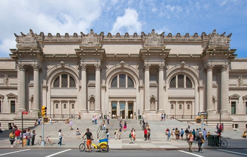 An exterior shot of The Metropolitan Museum of Art on a sunny summer day. Pedestrians walk along the sidewalk and some sit on the front stairs. A man with a yellow bike is in the street.