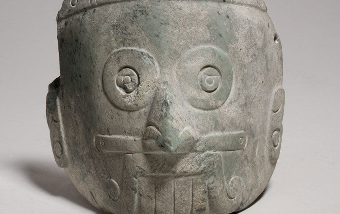 Rain god mask carved from stone with ringed eyes, prominent teeth, and a mouth with an upper lip-moustache that curls on each side