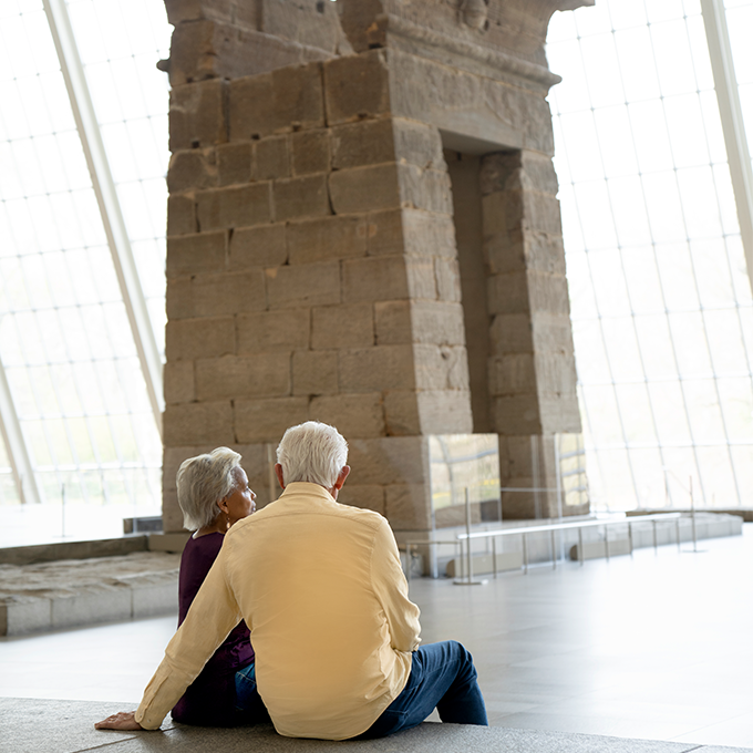 An older couple is seated with their backs turned towards us looking at the Temple of Dendur.