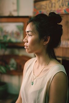Mei Lum stands in profile. She is stood against a blurred background of paintings and plants. Her demeanor is relaxed. Her dark hair is clipped on top of her head, and she is wearing a white, sleeveless blouse. A jade pendant hangs from her neck, against her chest.