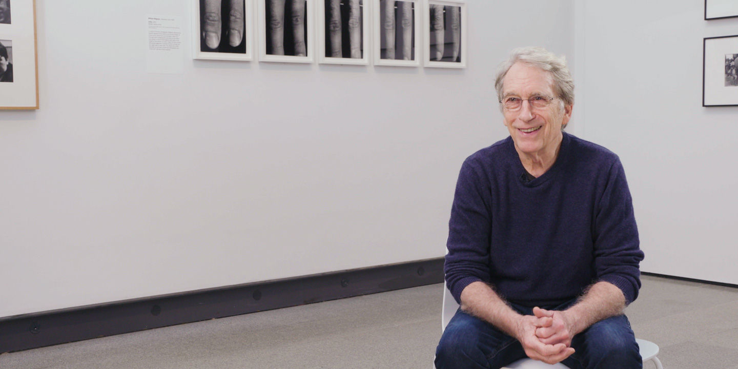 Video still of William Wegman seated and smiling during an interview in the exhibition Before/On/After