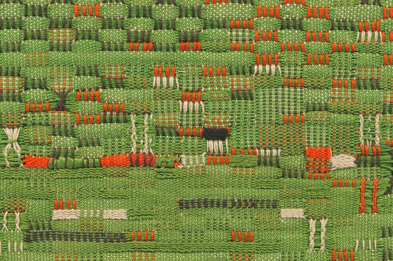 Detail of a woven material with grass green as the primary color, accented with dark green, oranges, and whites.
