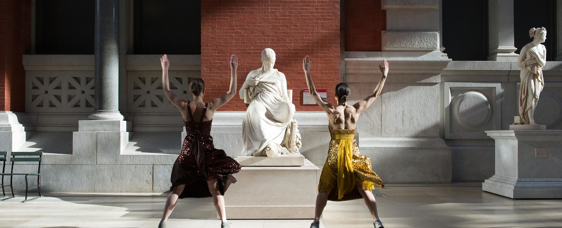 Two standing women, facing a statue of Sappho on opposites sides, with arms and legs spread during a museum workout