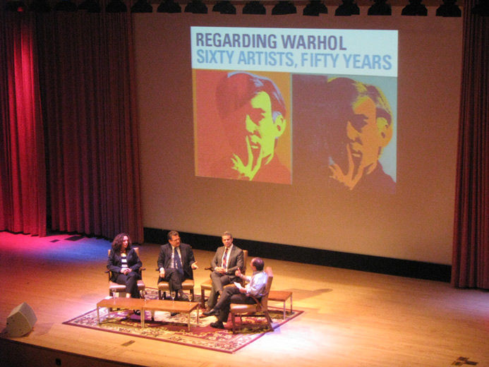 An elevated close-up of the stage of large modern blonde wood auditorium as viewed from the balcony; on stage is four people in low, comfortable chairs around a low table, behind them projected on screen is the title screen of the lecture: 