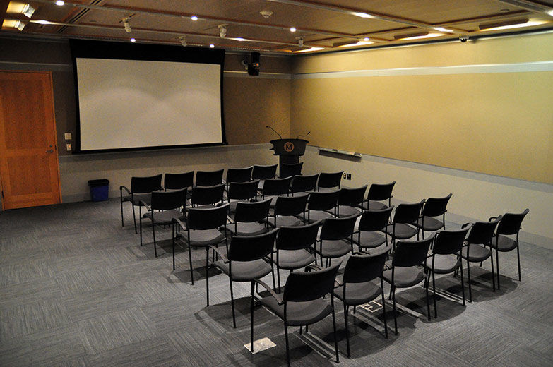A small, comfortable, modern, carpeted room with gray fabricwalls and gray paneling; the room is set with rows of chairs facing a projection screen and lectern 