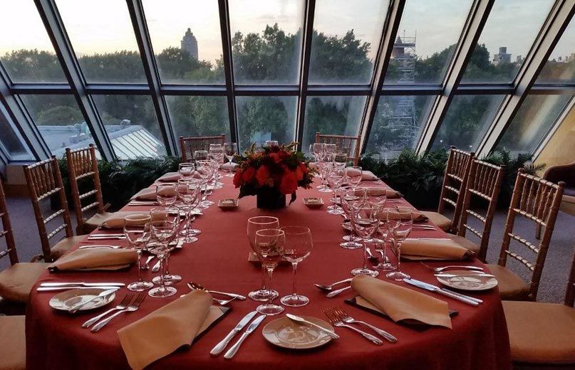 An intimate and elegant glass-enclosed room; a dining room table covered in a dark red table cloth is set with an elegant yet casual service and a low, elegant flower arrangement