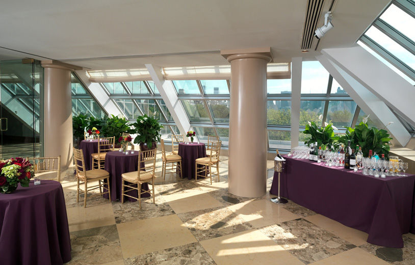 A bright, sunlit interior terrace set with small cafe tables draped in dark purple table cloths, and small, colorful flower arrangments; a large rectangular table is set with glass and beverages