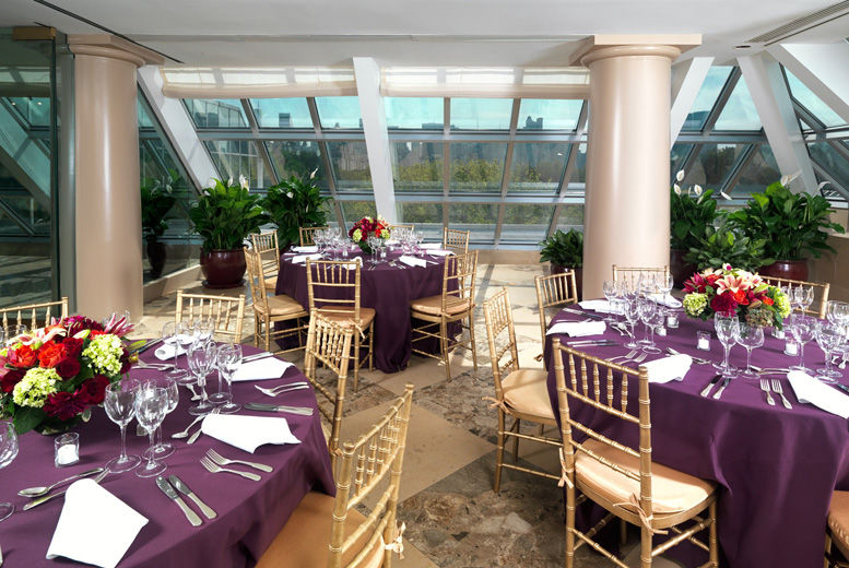 A bright, sunlit interior terrace set with round dining tables draped in dark purple table cloths and casual table service