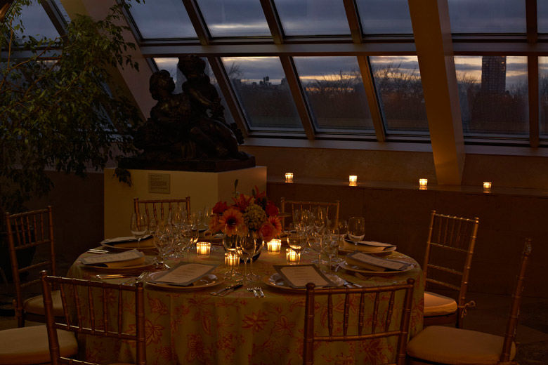 A intimate, dimly lit, glass-enclosed dining room with a round dining table set with a formal service, votive candles, and a low, colorful flower arrangement
