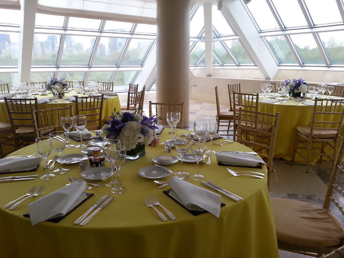 A bright, sunlit interior terrace set with round dining tables draped in bright yellow table cloths, a casual table service and large, elegant purple and white flower arrangments