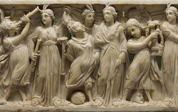 A white stone high relief carved sarcophogus depicting a group of women in classical dress holding various artistic or musical instruments, triumphing over three women with mermaid tails and wings, who have been pushed to the ground