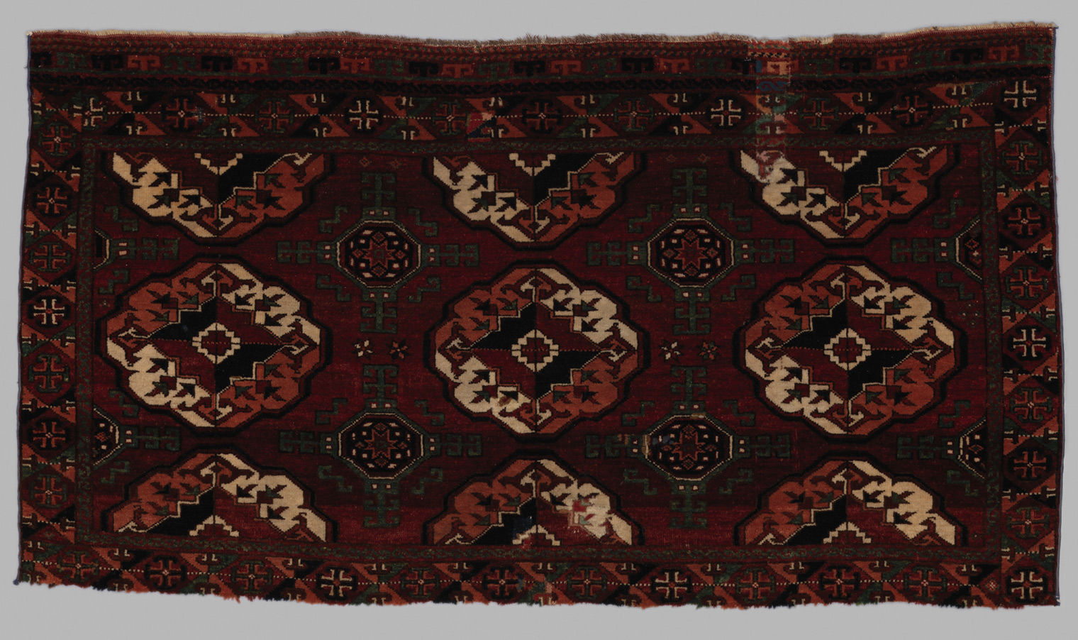 A close-up of a dark brown, burgundy, olive-green and white carpet decorated in a repeating pattern of geometric motifs