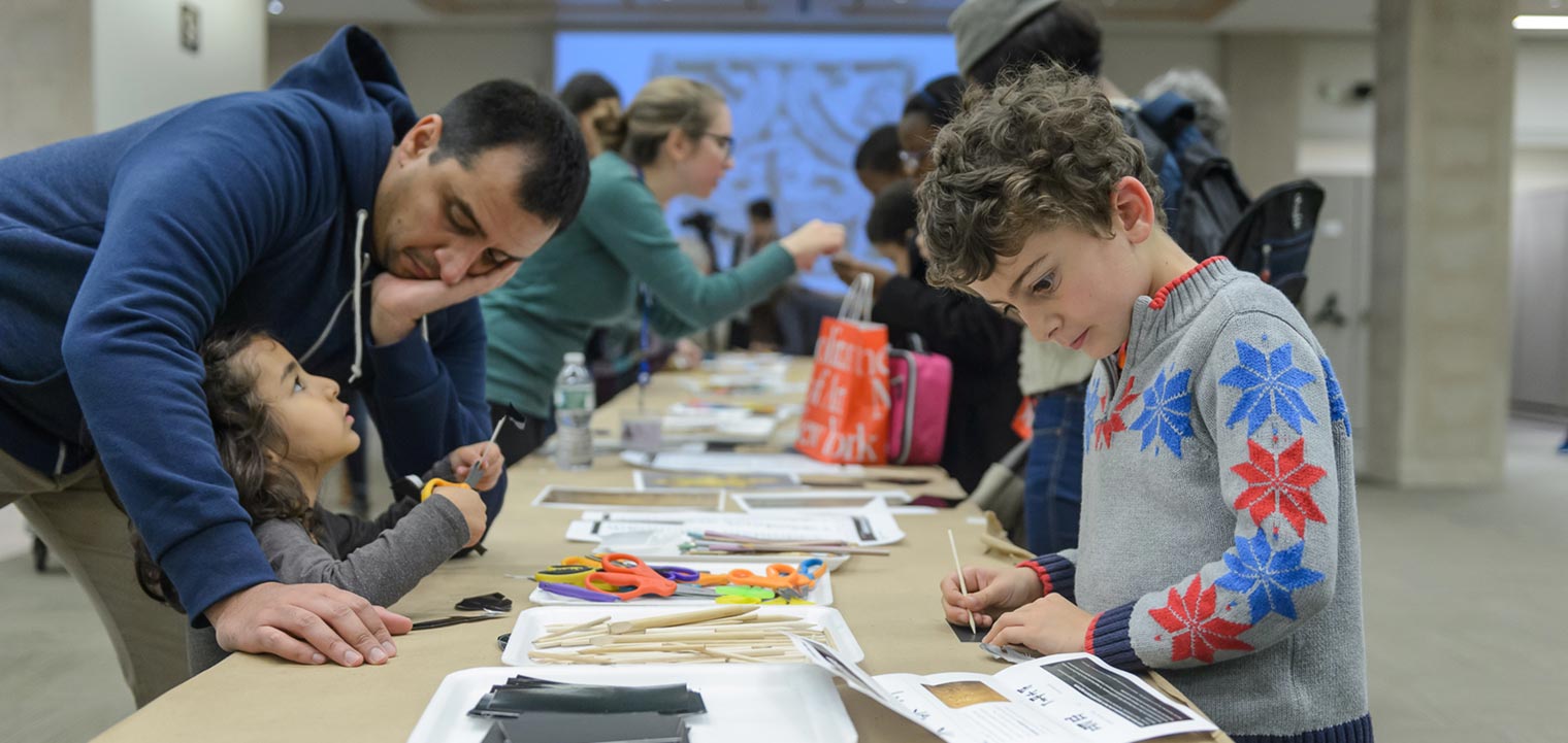 A long table covered with crafts projects with one boy writing on a paper and a young girl looks up to her father 