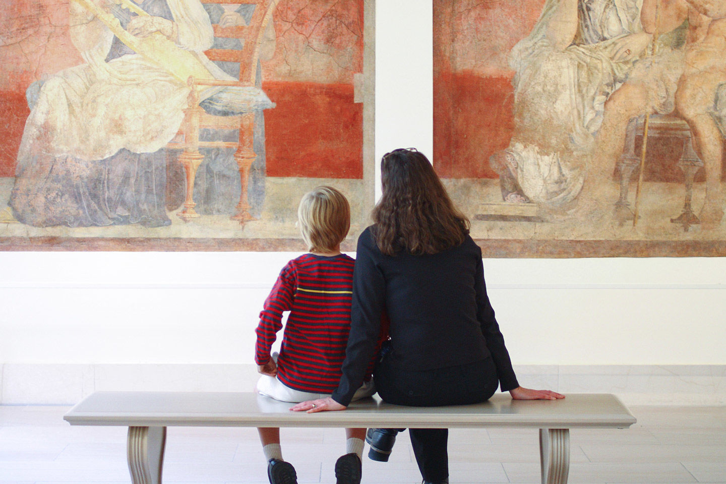 A woman and a child sit close together on a bench in a gallery looking at frescoes
