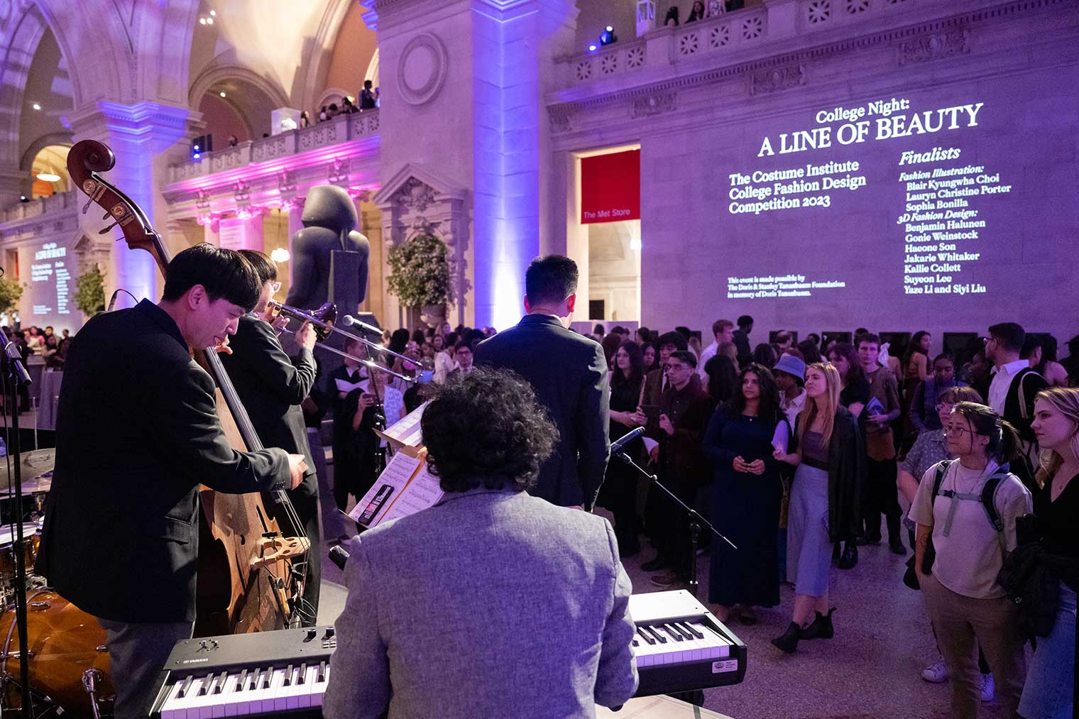 A keyboard player wearing a gray blazer and a double bass player perform music to a crowd of young people in a museum gallery.