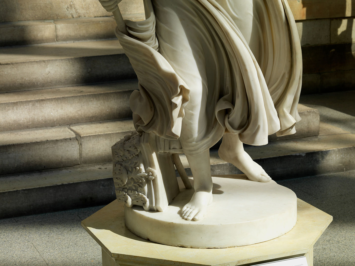 An image of the almost life-sized marble white sculpture Nydia, the blind flower girl of Pompeii, of her lower half and her flowing drapery taken in the American Wing Engelhard Sculpture Court at The Met, a skylit space with direct, dramatic natural light. Her feet are visible at the round base of the sculpture, along with a fallen Corinthian column and her staff along her right food. There are steps in the background of the shot. 
