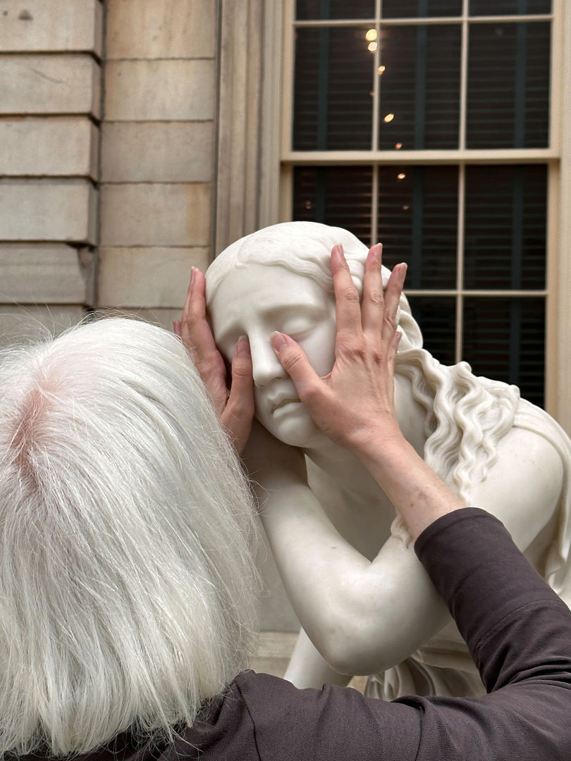 Image of the writer and professor Georgina Kleege, with her hands around the head of Nydia, The Blind Flower Girl of Pompeii, during a touch tour in the American Wing Engelhard Sculpture Court at The Met, a skylit space with direct natural light and the Façade of a 19th-century bank can be seen in the background. The image shows the back of the head of a grey-white-haired woman with medium long straight hair with a long-sleeved brownish purple sweater touching the face of an almost life-sized white marble statue of a young girl with her stretched-out hands, palms on the cheeks, and thumbs on the eyes and nose.